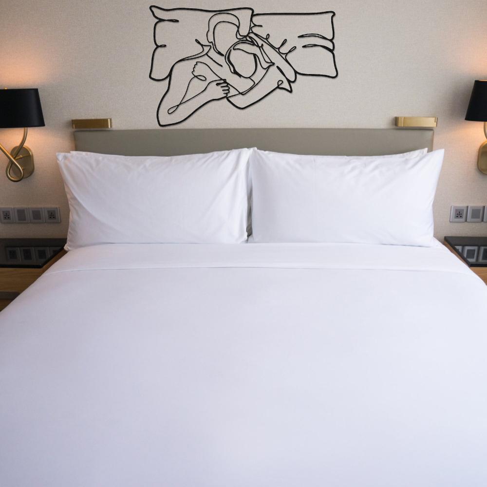 Abstract metal wall art of a couple embracing, mounted above a neatly made hotel-style bed with white linens, flanked by modern bedside lamps.