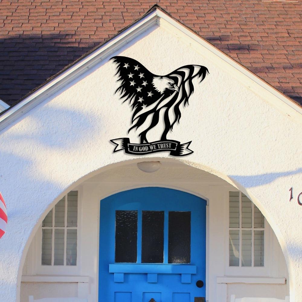 Black metal wall art of an American bald eagle with the U.S. flag and "In God We Trust" text, displayed on the exterior wall of a house above a blue front door with an arched entryway