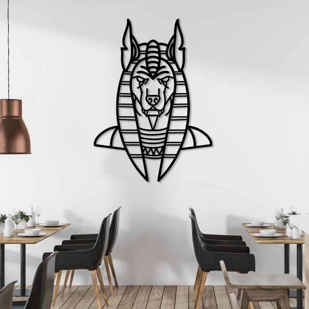 Black metal wall art of Anubis, the ancient Egyptian god, with a geometric design, mounted on a white wall in a modern dining setting. The space features wooden tables, black chairs, and elegant table settings, complemented by a bronze pendant light.