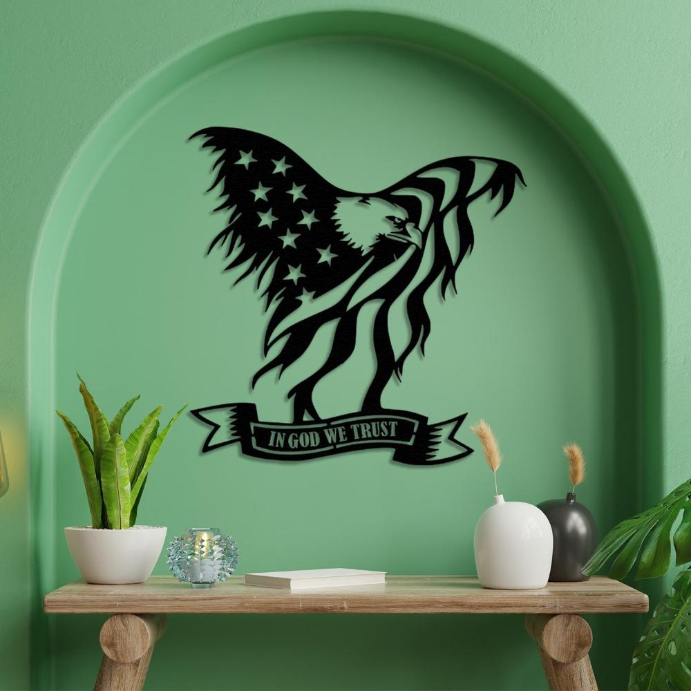 Black metal wall decor featuring a bald eagle with an American flag design integrated into its wings and the text "In God We Trust" displayed on a ribbon below, mounted on a green arched wall above a rustic table with decorative plants.