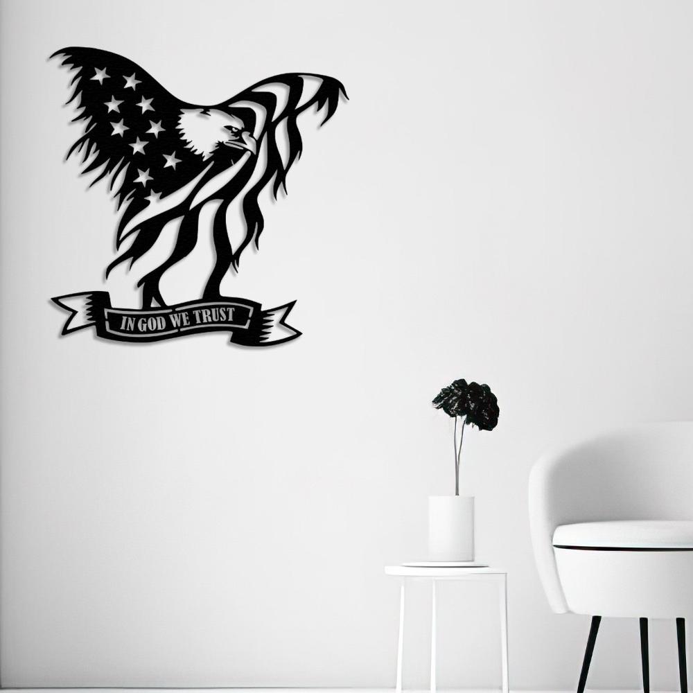 Black metal wall art of an American bald eagle holding the U.S. flag with the text "In God We Trust," mounted on a minimalist white wall above a sleek white chair and a small table with a black flower in a vase.