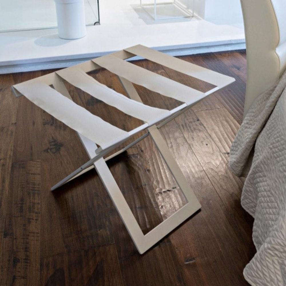 A modern beige metal luggage rack with sturdy fabric straps, placed on dark wooden flooring next to a cream-colored chair, in a minimalist bedroom setup.