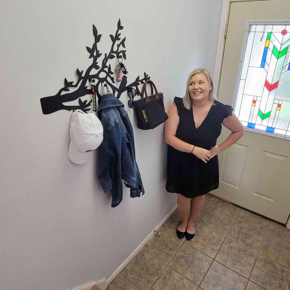 A woman stands near a black metal wall-mounted coat rack with bird and branch designs in an entryway. The rack holds a white cap, a denim jacket, a black handbag, and keys, providing a functional and stylish solution for organizing personal items near the door.