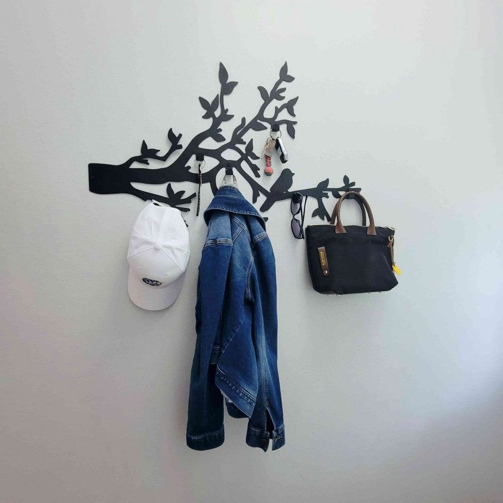 A black metal wall-mounted rack with bird and branch designs, used as an entryway organizer. The rack holds a white cap, a denim jacket, a black handbag, sunglasses, and keys, providing a functional and stylish solution for organizing personal items.