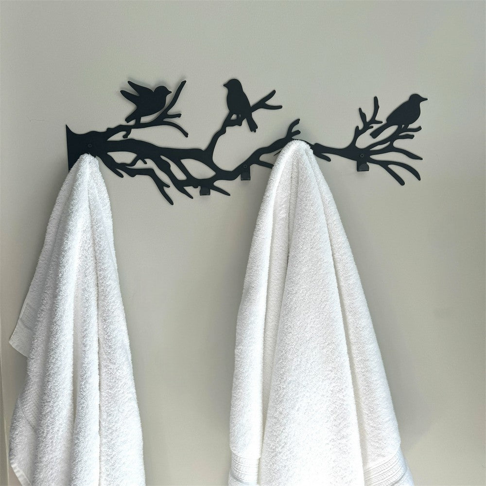 A black metal wall-mounted rack featuring birds on tree branches, used here as a towel holder. The design incorporates silhouettes of birds perched on branches, adding a touch of nature to the decor. Two white towels hang neatly from the hooks, making it a practical and decorative piece for bathrooms or bedrooms.