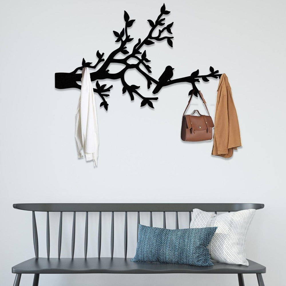 A black metal wall-mounted coat rack designed with birds and branches, holding a white shirt, a brown handbag, and a beige coat. Below the rack, a stylish grey bench with decorative pillows adds a modern touch to the entryway or hallway decor.