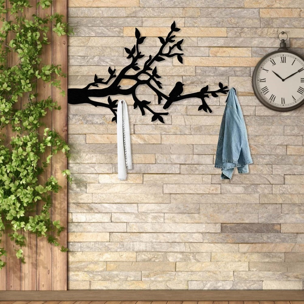 A black metal wall-mounted coat rack designed with bird and branch silhouettes, installed on a brick wall. The rack holds a white scarf and a denim jacket, with a large wall clock and green plants enhancing the rustic, natural decor.