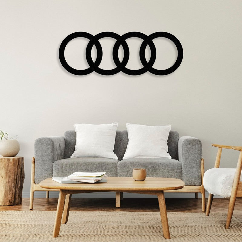 Black metal Audi logo wall art featuring four interlocked rings, mounted above a modern gray sofa with white cushions. The room is styled with a wooden coffee table, a natural stump side table, and minimalistic decor for a cozy and elegant ambiance.