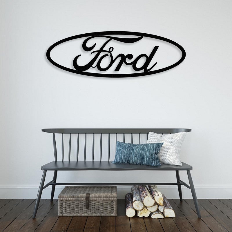 A stylish black Ford-inspired metal wall art mounted on a white wall above a sleek black bench adorned with decorative pillows. The scene includes rustic elements like stacked firewood and a woven basket below, adding a cozy touch.