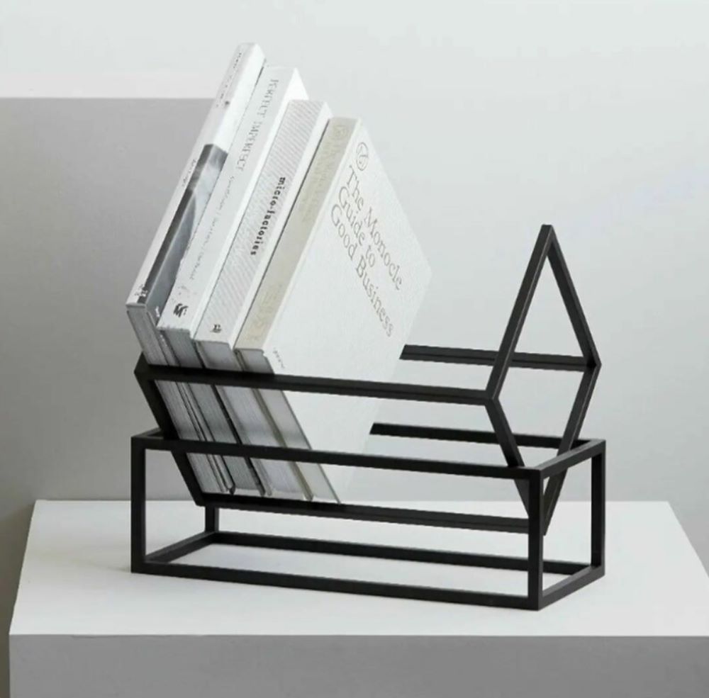 A black geometric book rack on a white pedestal, holding a selection of monochromatic books with a modern minimalist style