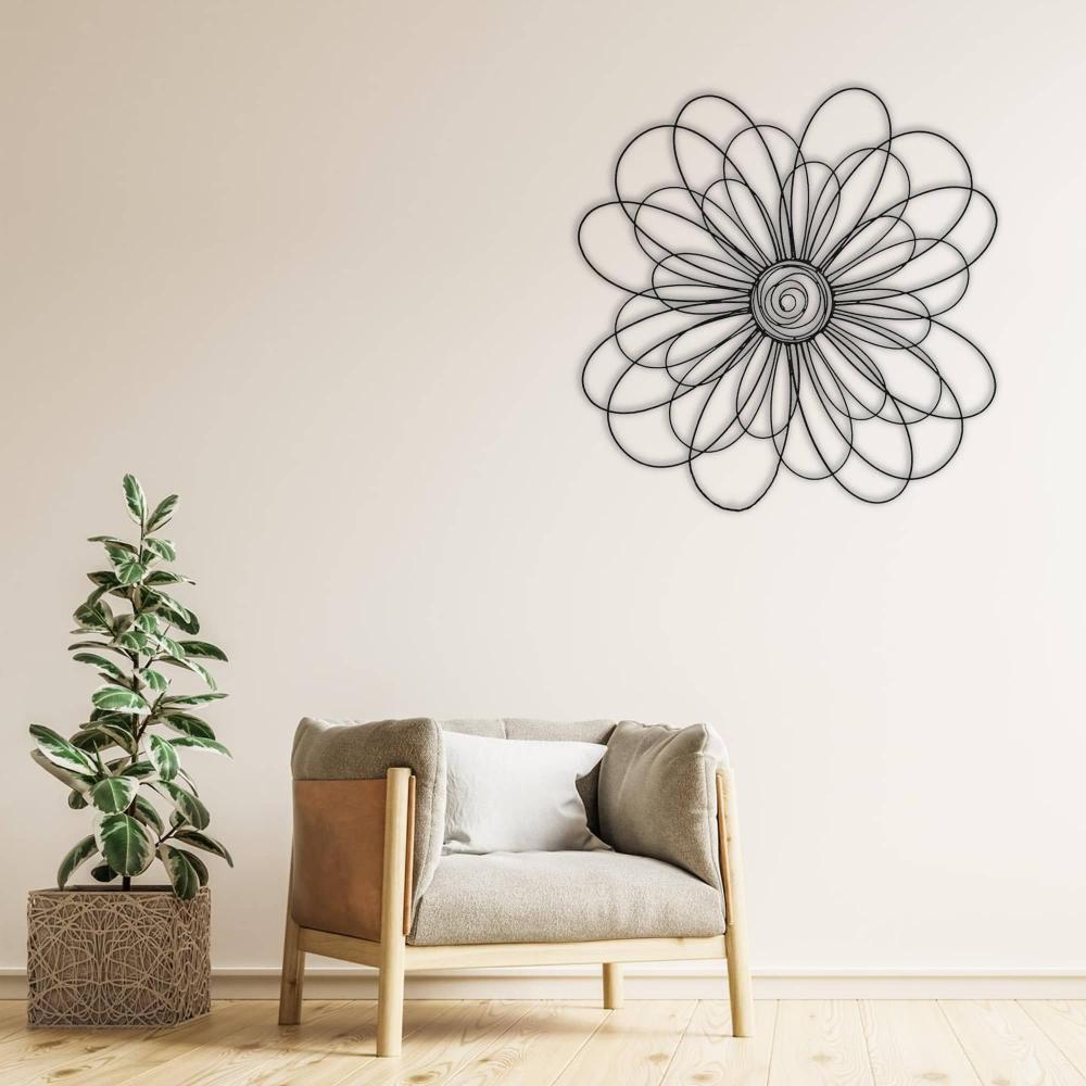 Black metal daisy wall art featuring a spiral center, mounted on a light beige wall above a modern beige armchair with a white cushion, next to a potted plant.