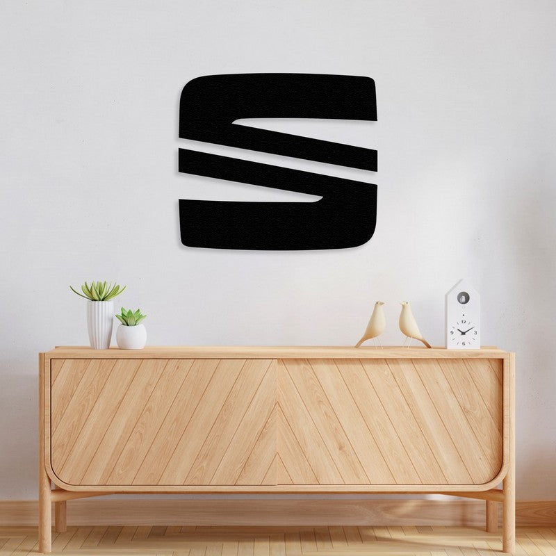 Black metal Seat logo wall art mounted on a minimalist white wall, placed above a light wooden console with a chevron pattern. The console is decorated with small potted plants, ceramic bird figurines, and a modern white clock, creating a clean and contemporary look.