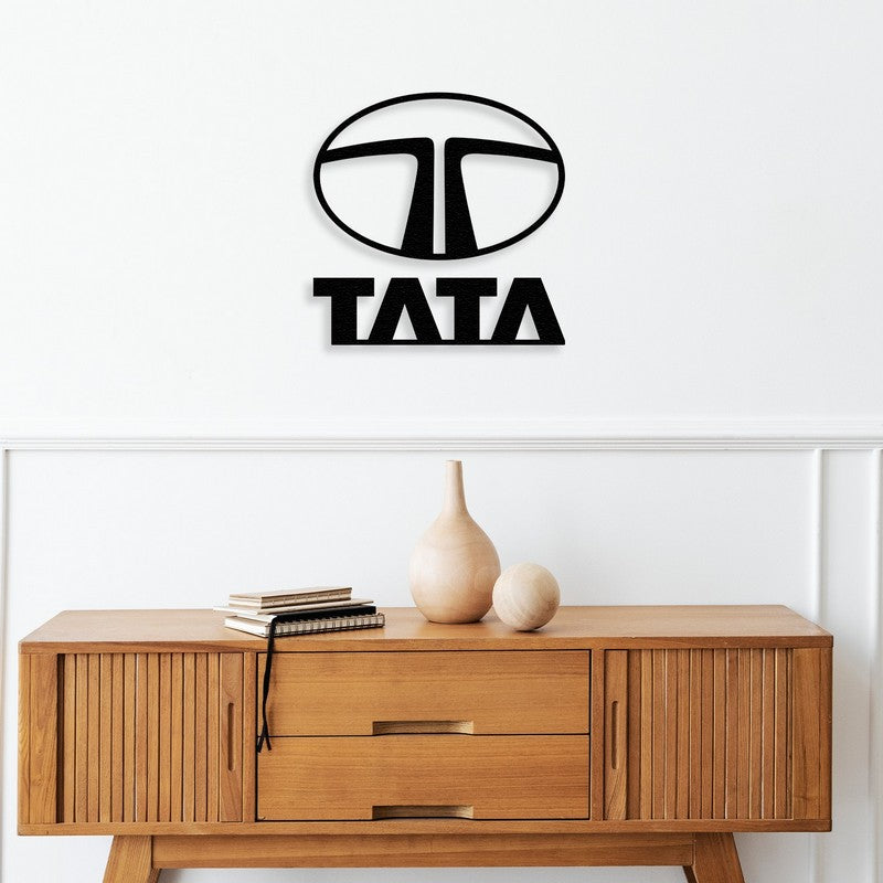 Black metal Tata logo wall art mounted above a mid-century modern wooden console table featuring clean lines and minimalist design. The console is adorned with wooden vases and stacked books, adding a natural and sophisticated touch to the decor.