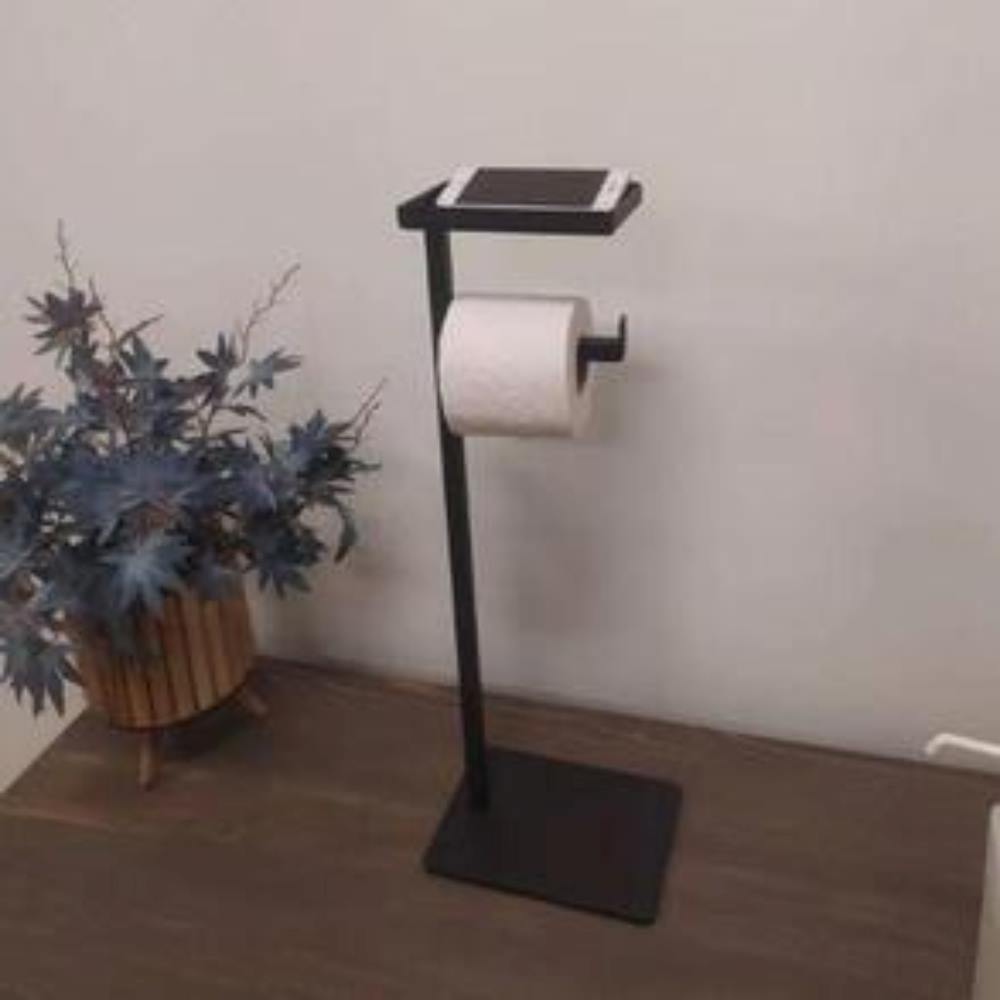 A black floor-mounted toilet paper holder with a compact shelf holding a smartphone, placed on a wooden surface next to a decorative plant.