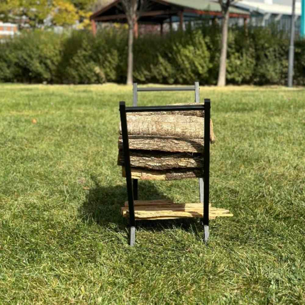 Front-facing view of a black V-shaped wood rack outdoors on lush green grass, holding an organized stack of firewood with a lower compartment for smaller logs.