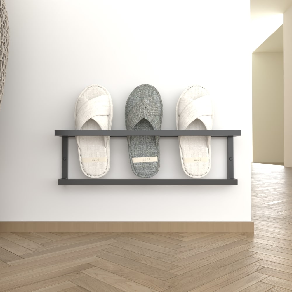 A modern black wall-mounted slipper rack neatly displaying three pairs of slippers, positioned on a light wall with herringbone wooden flooring for a contemporary look.