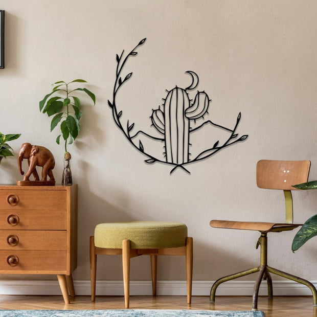 Black metal wall art featuring a stylized cactus with a crescent moon and leaf design, mounted on a light-colored wall. Below is a wooden dresser with a carved elephant figurine, a green cushioned stool, and a wooden chair with a vintage aesthetic.