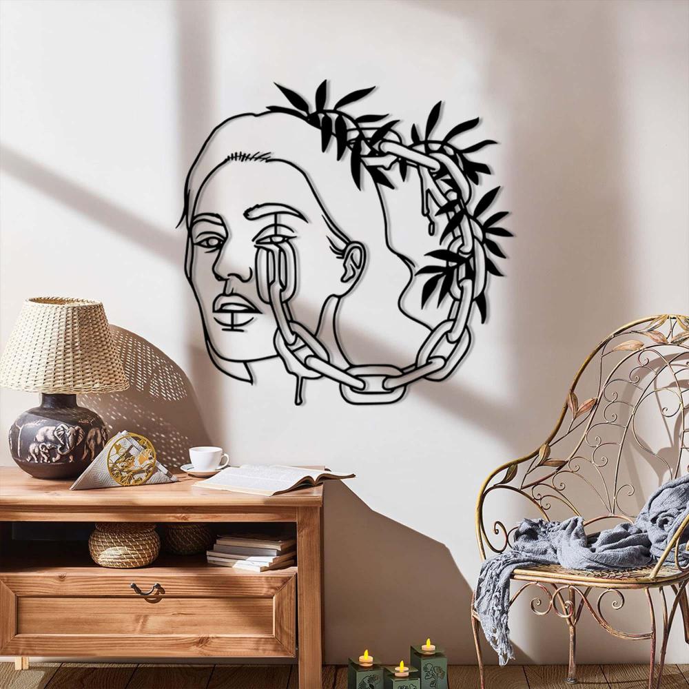 A bright corner with black metal wall art of a woman's face adorned with chains and leaves, mounted above a wooden console table and decorative items.