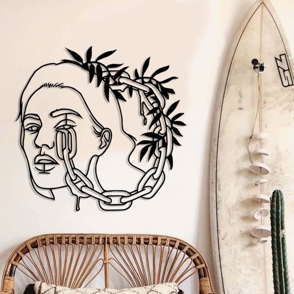 A minimalist interior with a black metal wall art of a woman's face entwined with chains and leaves, mounted next to a surfboard and a wicker chair.