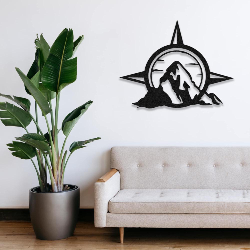 Metal wall art featuring a compass and mountain design placed above a beige sofa with a large green plant in a minimalist living room.