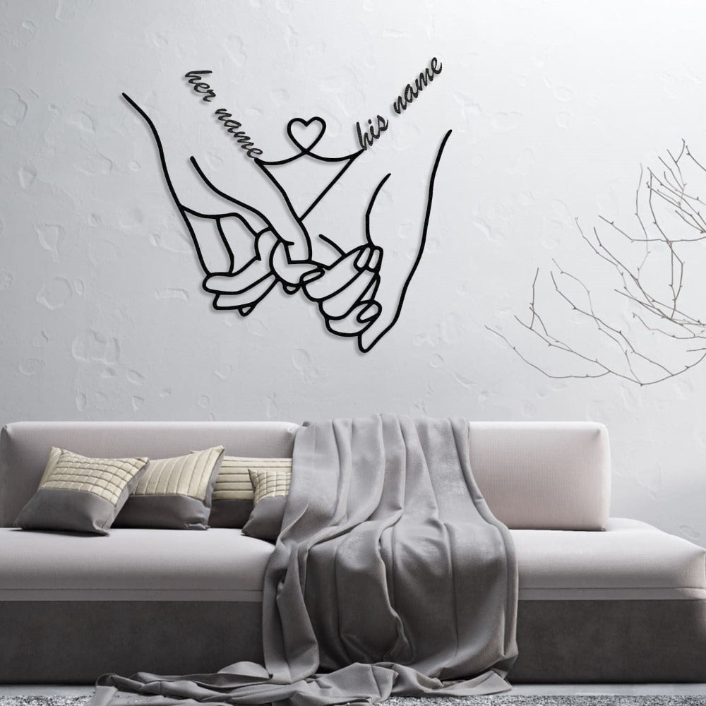 Personalized metal wall art featuring intertwined hands with "her name" and "his name" elegantly connected by a heart, displayed on a textured white wall above a modern sofa with gray cushions and a matching throw.