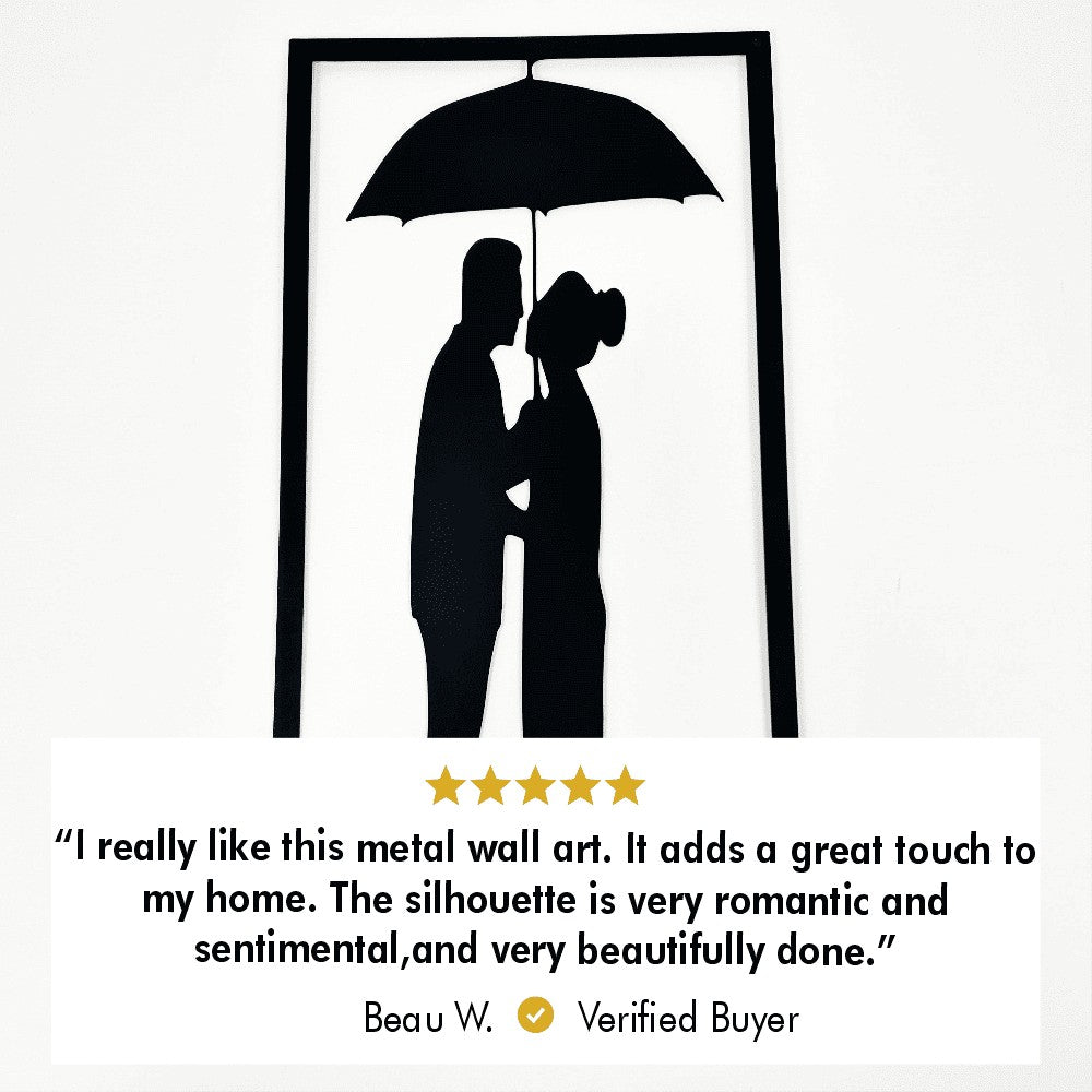 Close-up of a black framed metal wall art depicting a couple under an umbrella. A customer review beneath the artwork reads: "I really like this metal wall art. It adds a great touch to my home. The silhouette is very romantic and sentimental, and very beautifully done." The review is from Beau W., a verified buyer.