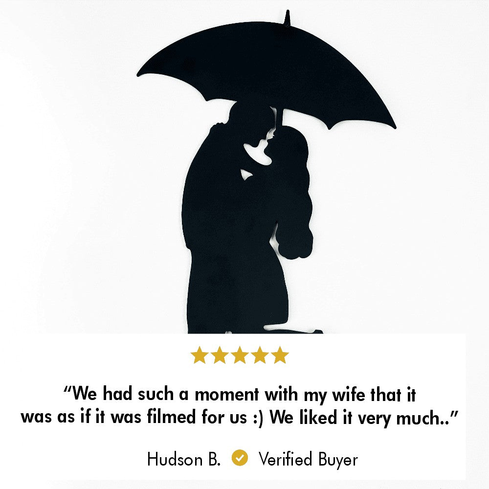 A close-up view of a black metal wall art featuring a couple embracing under an umbrella. Below the art is a verified customer review from Hudson B., who describes a heartfelt experience with the piece, saying it felt as though the moment was filmed just for them, expressing how much they enjoyed it.