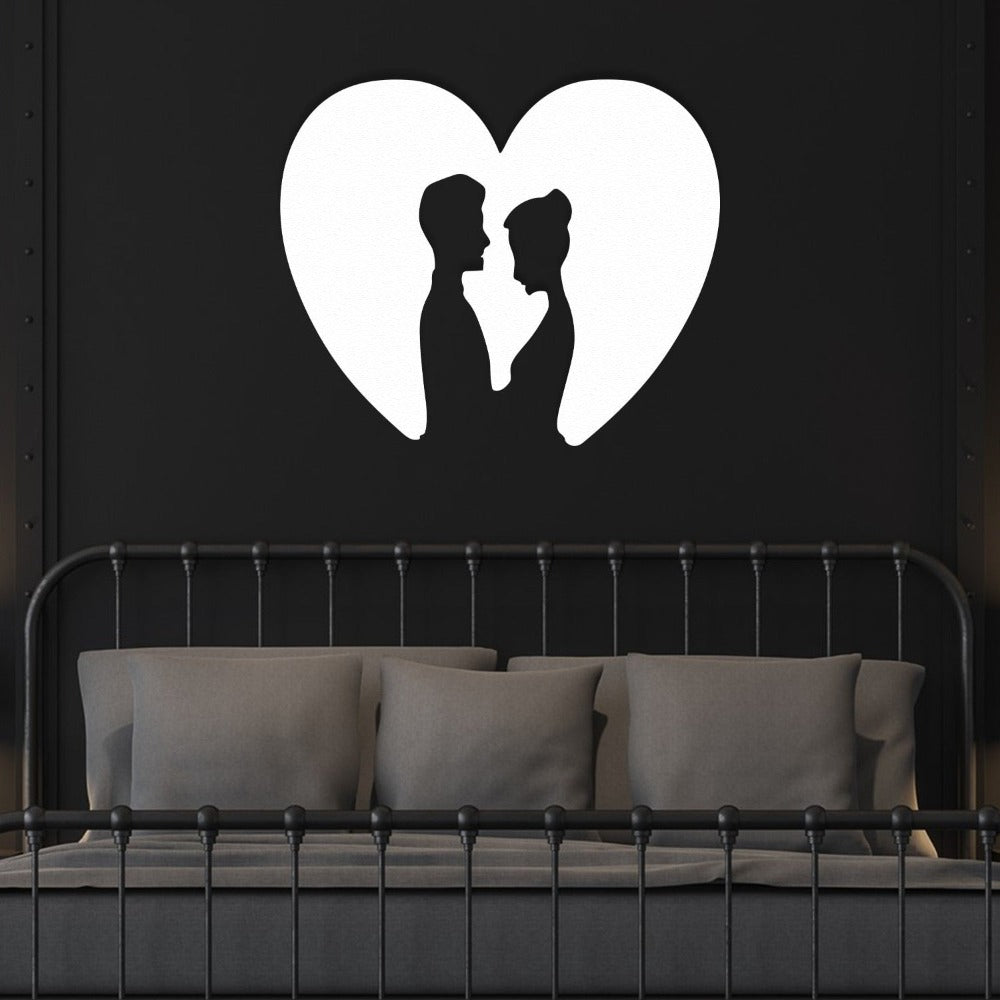 White metal wall art of a couple inside a heart silhouette, mounted on a dark wall above a bed in a modern bedroom with monochrome accents.