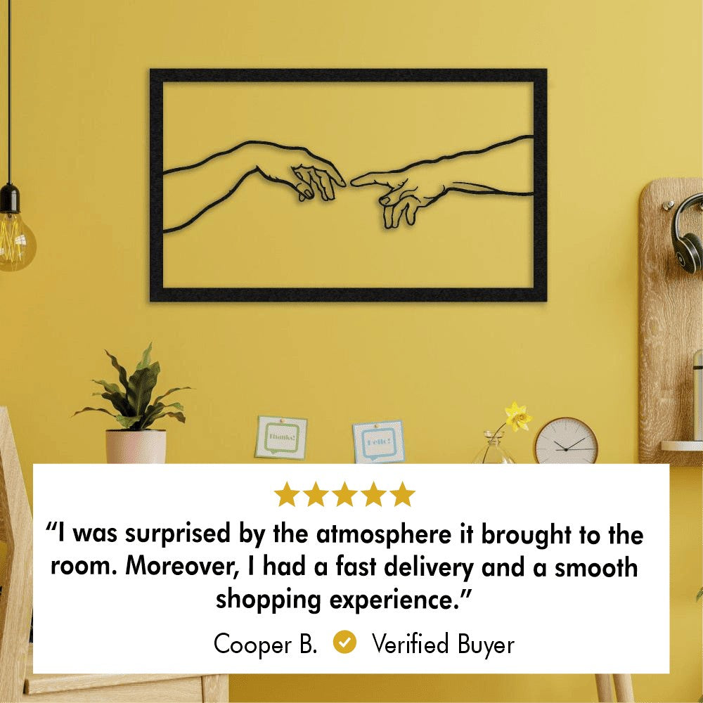 Metal wall art inspired by Michelangelo's "The Creation of Adam," featuring two hands reaching towards each other, framed and mounted on a bright yellow wall. Below the artwork is a 5-star customer review from a verified buyer, Cooper B., stating: "I was surprised by the atmosphere it brought to the room. Moreover, I had a fast delivery and a smooth shopping experience."