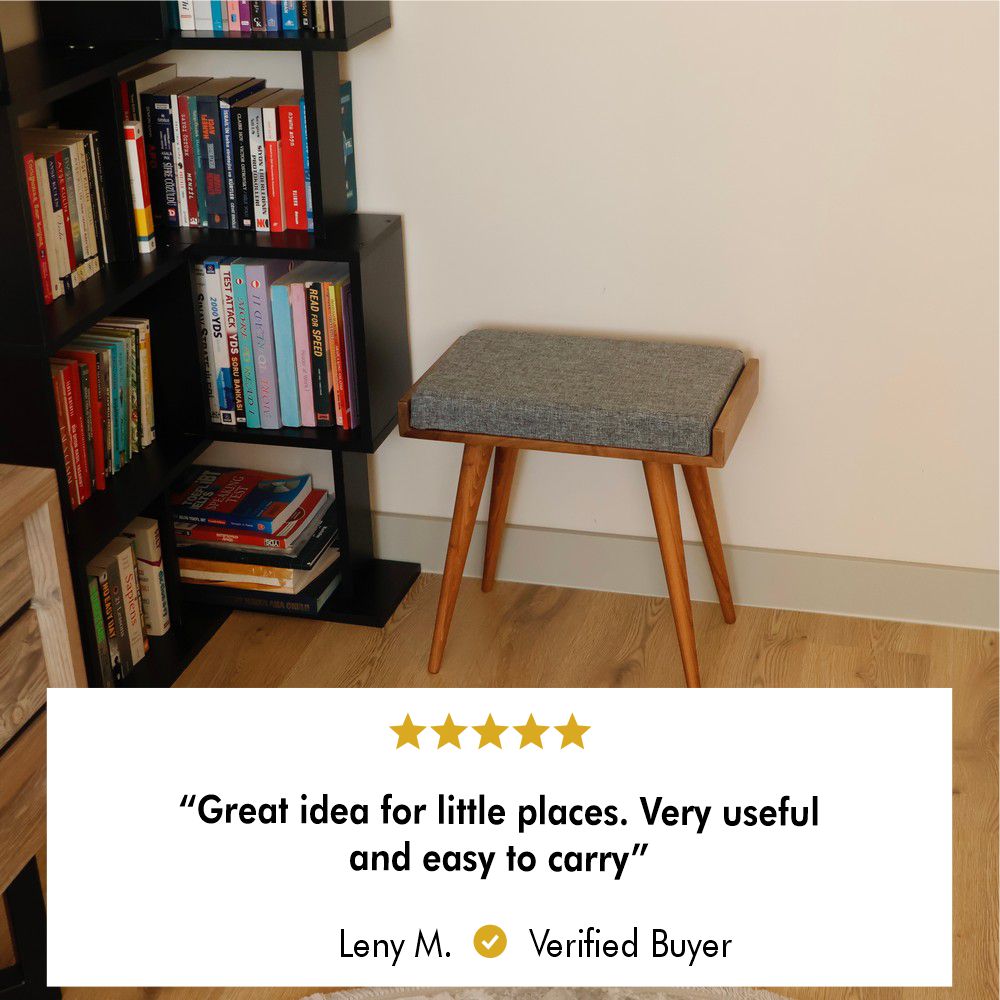 A wooden stool with a grey cushion seat positioned next to a black bookshelf in a cozy reading corner.