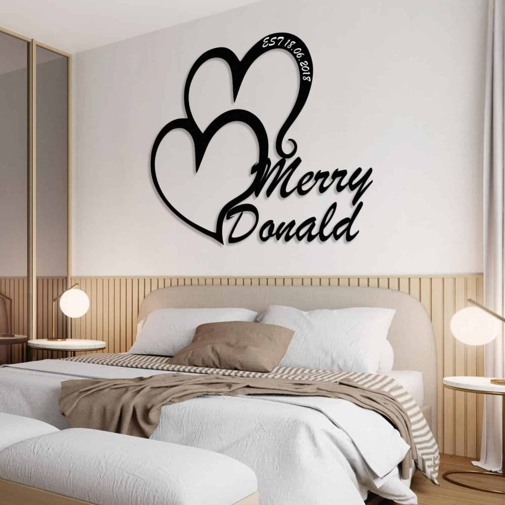 Custom double-heart metal wall art with the names "Merry" and "Donald," displayed above a cozy, modern bedroom setup with wooden panel accents, neutral bedding, and bedside lamps.