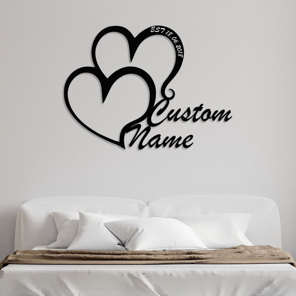 Customizable double-heart metal wall art featuring "Custom Name" displayed above a minimalist white bed with neutral pillows and a cozy throw blanket.