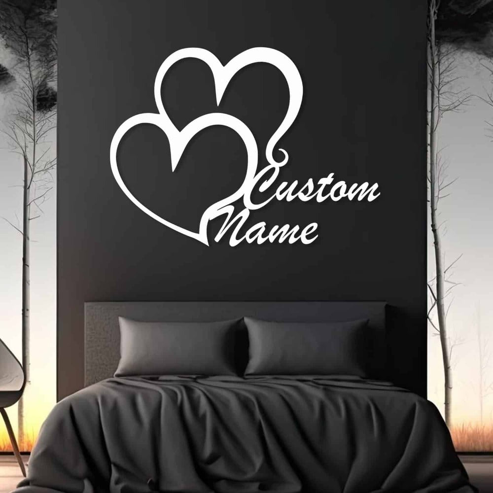 White double heart metal wall art with customizable names displayed on a dark wall in a modern bedroom setting, featuring a black bedspread and an abstract forest design in the background.