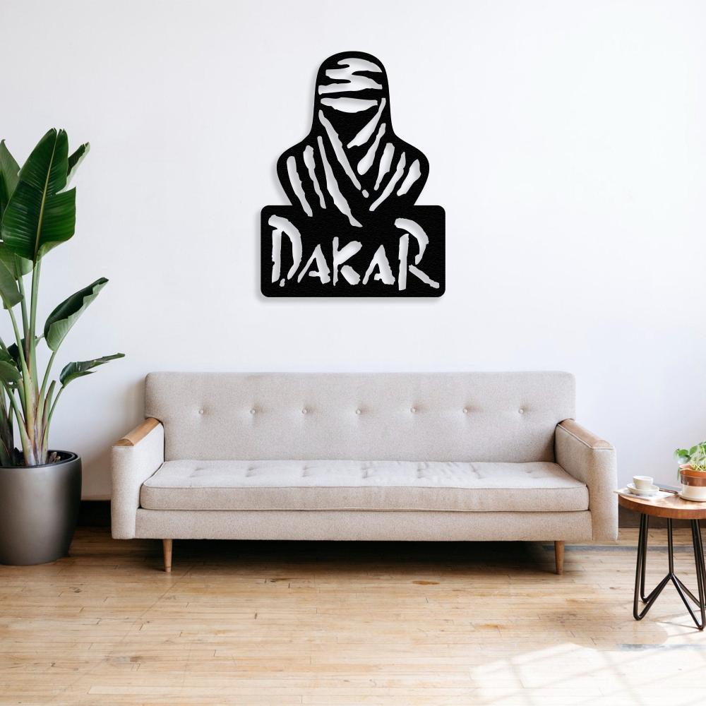 Black Dakar Rally metal wall art featuring a silhouette of a desert nomad and bold "Dakar" text, mounted on a white wall above a minimalist beige tufted sofa with wooden arms. The room includes a green potted plant on the left and a wooden round side table with decor items on the right, placed on a light wooden floor.