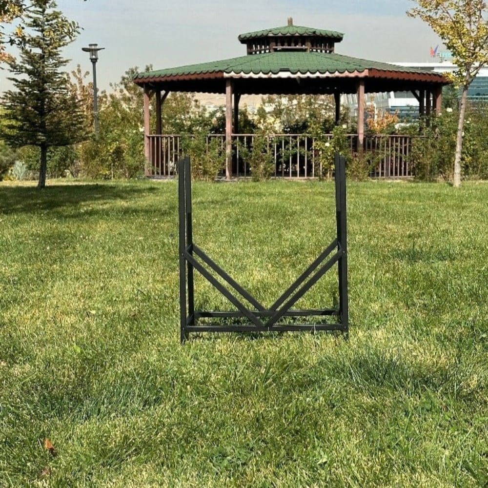 An empty geometric firewood rack in black metal, with a V-shaped design, placed outdoors on green grass, with a scenic view of a gazebo in the background