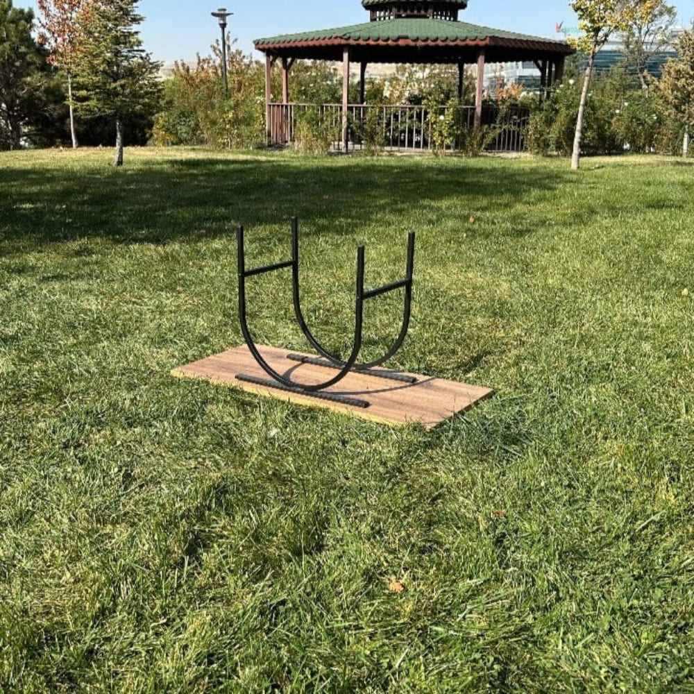 A black oval-shaped metal log holder without logs, placed on a wooden plank in an outdoor park setting with green grass and a gazebo in the background