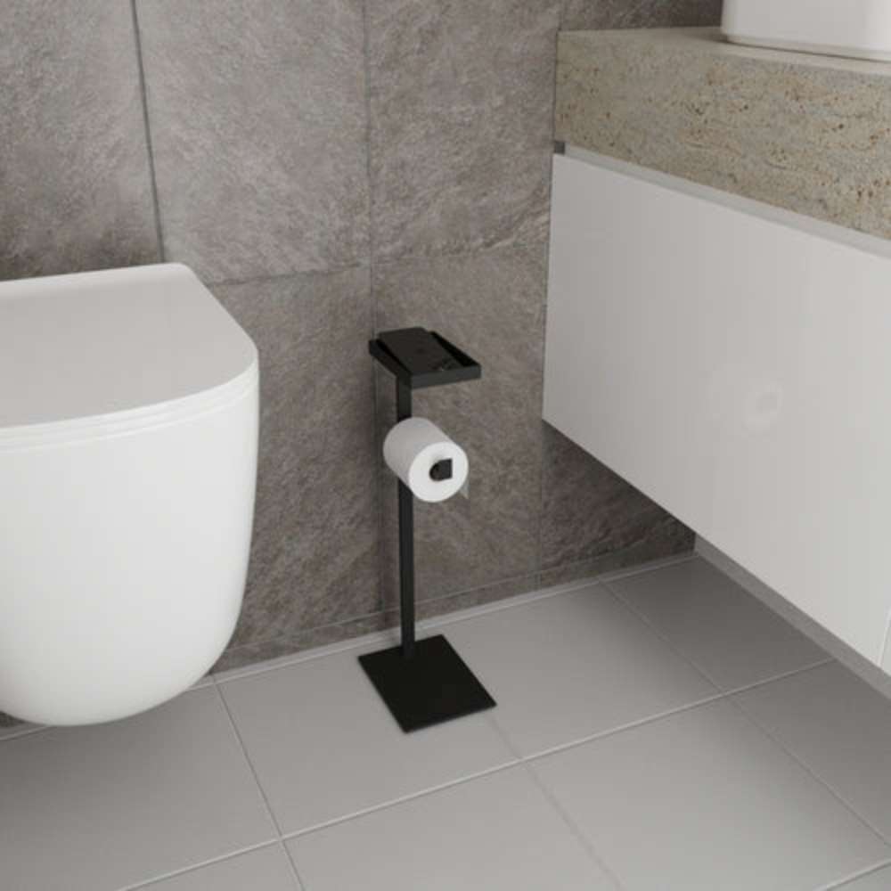 A sleek black floor-mounted toilet paper holder with a built-in shelf for small items, placed in a modern bathroom with gray tiled walls.