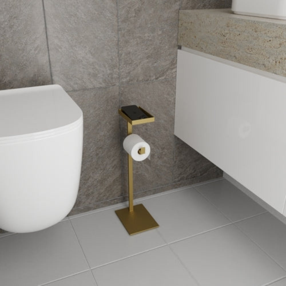 A gold floor-mounted toilet paper holder with a small shelf for accessories, displayed in a modern bathroom setting with tiled walls and sleek finishes.