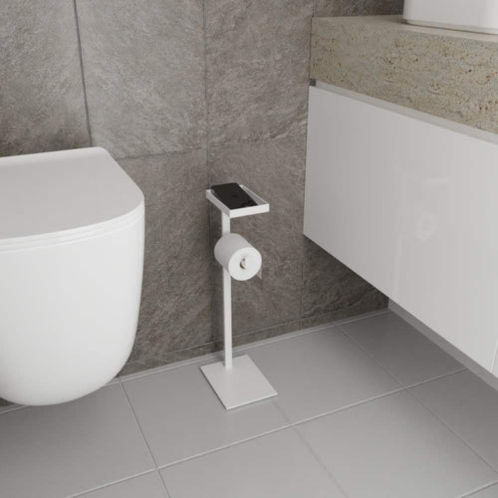 A white floor-mounted toilet paper holder with a built-in shelf, styled in a bathroom with light gray tiles and minimalist decor.