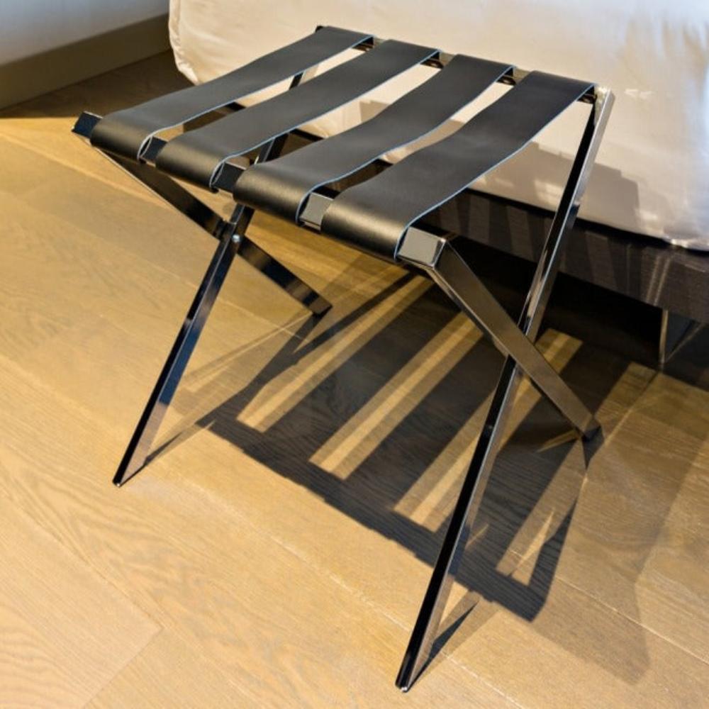 A stylish foldable metal luggage rack with genuine leather straps, placed on a wooden floor beside a bed, showcasing its contemporary design.