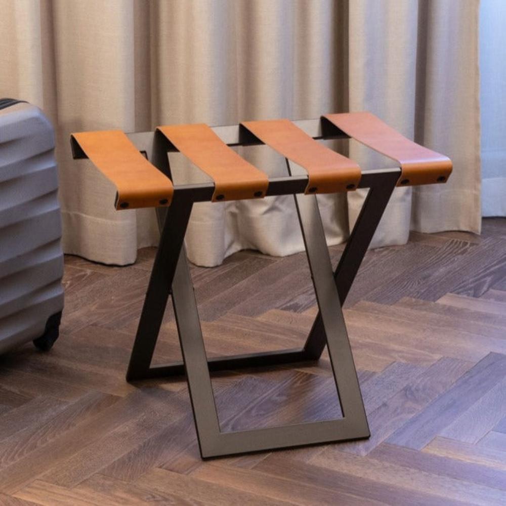 A foldable metal luggage rack with genuine leather straps, placed on a wooden floor in a room with beige curtains in the background.