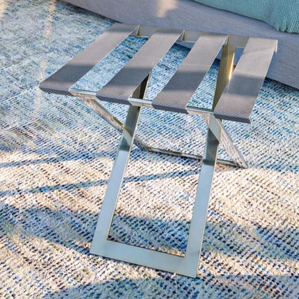 A foldable metal luggage rack with wide straps, displayed on a colorful textured rug next to a sofa, highlighting its portability and style.