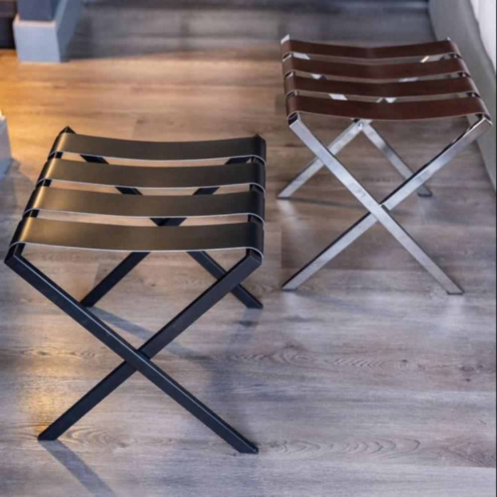 Two foldable luggage racks with leather straps, one in black and the other in chrome, illuminated by warm ambient lighting on a wood-textured floor.