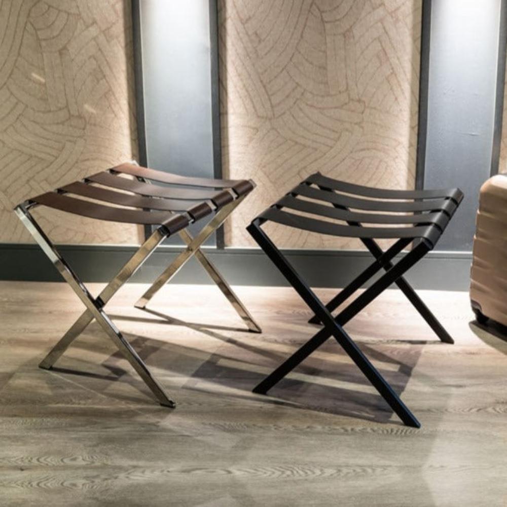 Two foldable metal luggage racks, one in black and the other with chrome legs and brown leather straps, side by side on wooden flooring in a modern interior.