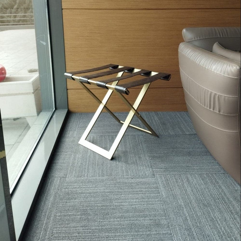 Foldable metal luggage rack with brown leather straps and a gold frame, placed near a large window in a modern seating area with gray carpet and beige leather furniture.