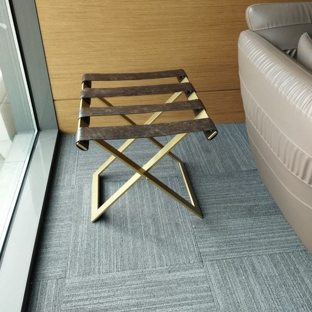 Foldable metal luggage rack with brown leather straps and a gold frame, placed in a modern seating area with beige leather chairs and gray carpet, providing a stylish and practical storage solution.