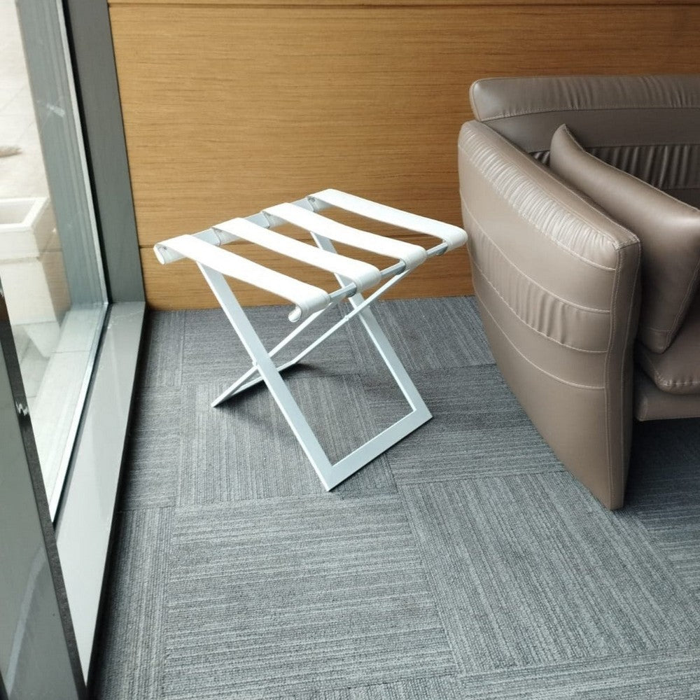 Foldable metal luggage rack with white leather straps, placed in a modern seating area with beige leather chairs and gray carpet, offering a sleek and functional design.