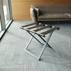 Foldable metal luggage rack with genuine leather straps, placed on a light gray carpeted floor near a modern leather sofa and a large window in a contemporary room.