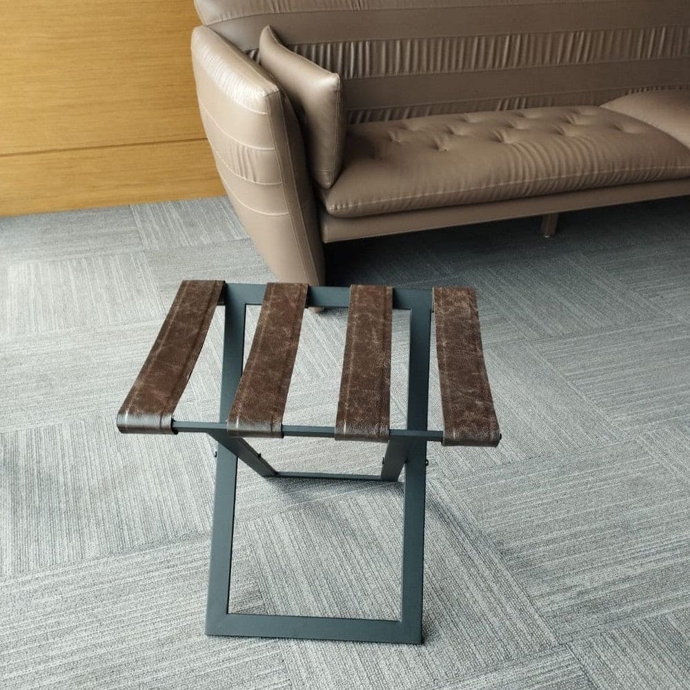 Foldable metal luggage rack with brown leather straps and a dark metal frame, placed in a modern seating area with beige leather furniture and gray carpet, providing a practical and stylish solution for luggage storage.