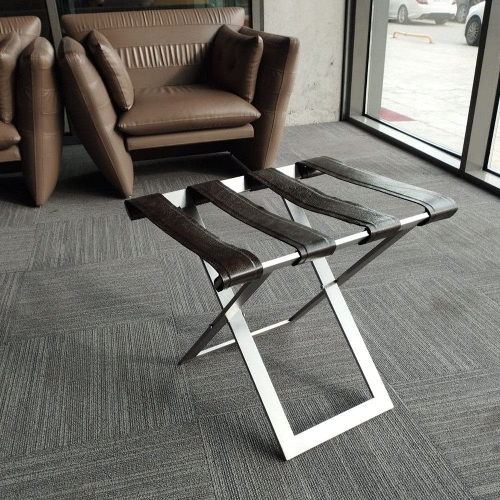 Foldable metal luggage rack with dark brown leather straps and a sleek metal frame, placed in a modern seating area with brown leather armchairs and gray carpet, offering a stylish and functional solution for luggage storage.